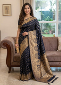 A Woman in a navy blue color banarasi silk saree with gold zari details standing in a living room.