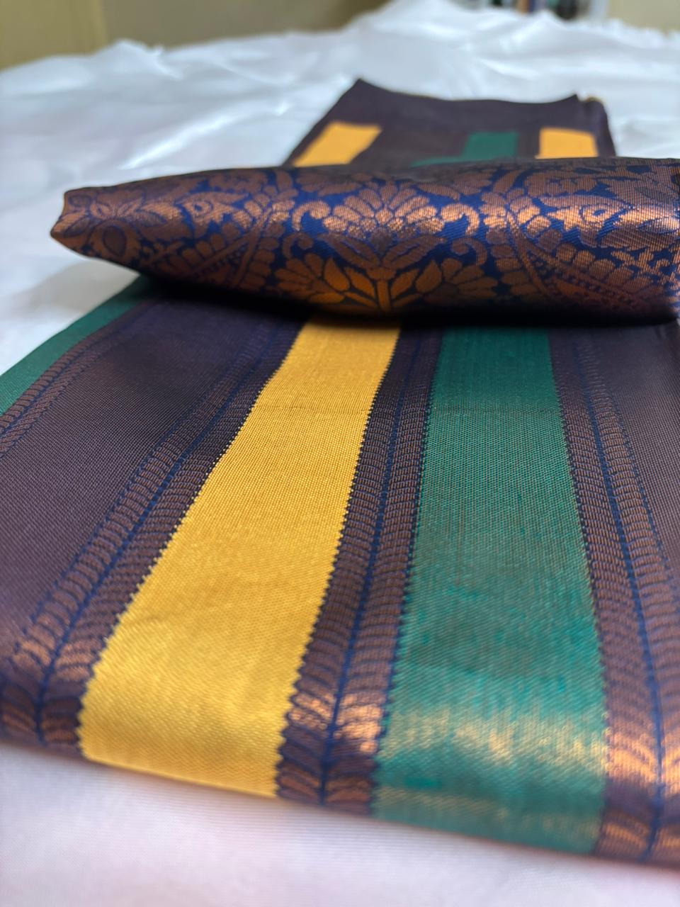 A folded silk saree with a metallic gold brocade floral pattern, featuring wide stripes of mustard yellow and emerald green on a dark purple base, displayed on a white surface.