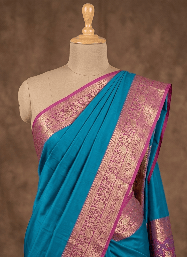 A turquoise blue silk saree with a wide pink and gold zari border, draped on a beige mannequin torso against a textured brown background.
