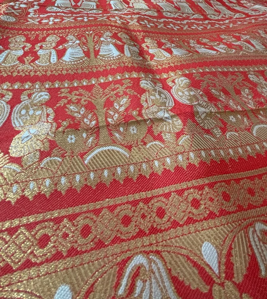 A close-up of a red and gold patterned pallu of a Baluchari Saree featuring intricate woven motifs of figures, trees, and ornate borders.