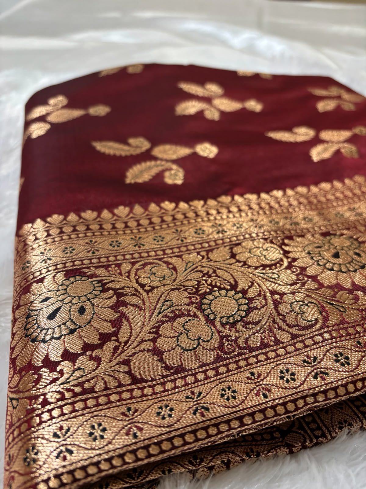 A folded deep maroon red silk saree Banarasi Saree with an intricate gold zari brocade border and floral motifs