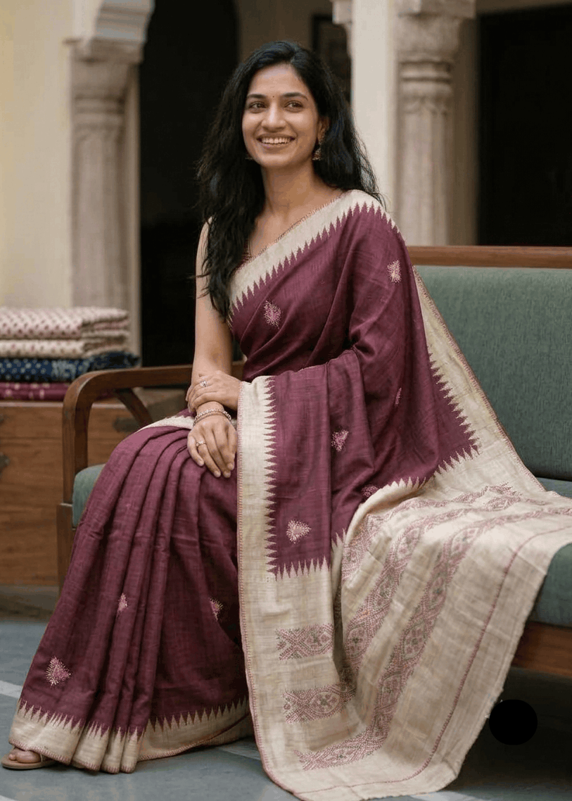A woman wearing a deep maroon Boolian Kantha saree with a beige border and small floral motifs, seated on a green sofa in an indoor setting.
