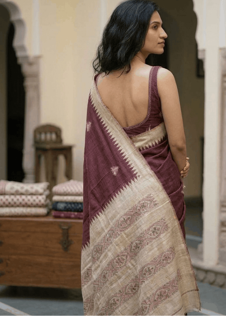 A woman wearing a deep maroon and beige patterned Boolian kantha saree, photographed from behind in an elegant courtyard with arches.