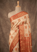 A cream-colored silk Baluchari saree with intricate red and gold zari patterns, draped on a mannequin against a textured brown backdrop.