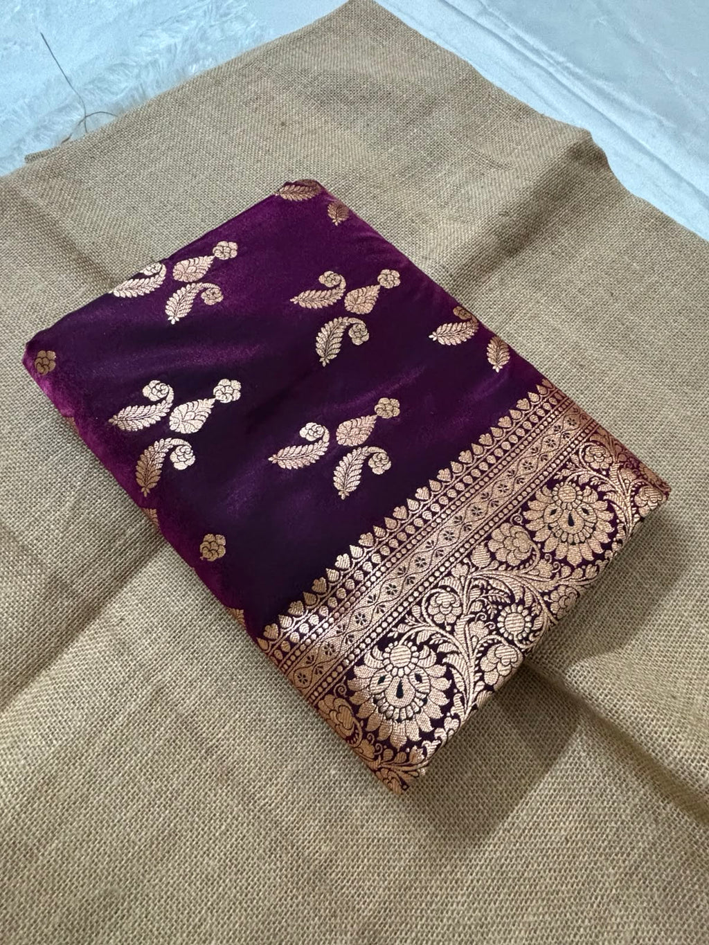 A folded deep Wine purple dual-tone silk Banarasi saree with intricate gold Banarasi zari embroidery and a wide ornate border, displayed on a textured beige burlap cloth.