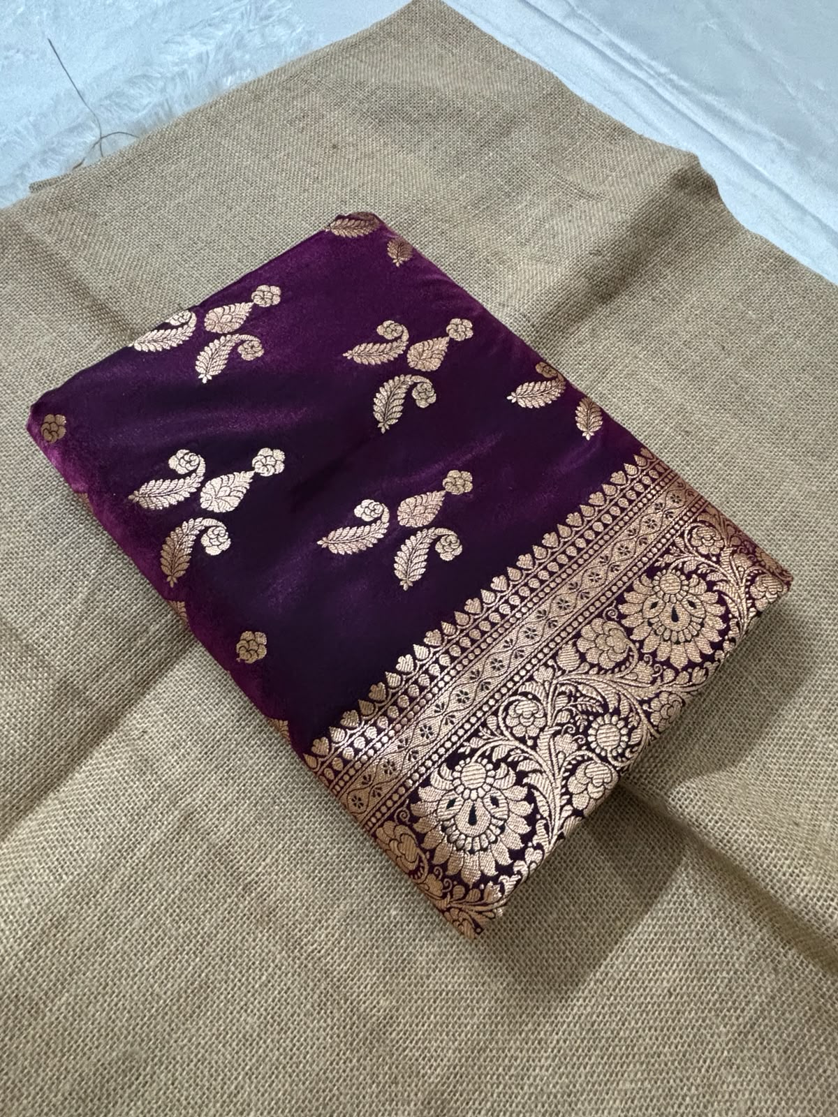A folded deep Wine purple dual-tone silk Banarasi saree with intricate gold Banarasi zari embroidery and a wide ornate border, displayed on a textured beige burlap cloth.