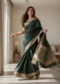 A woman wearing a dark green Banarasi silk saree with gold Zari work patterns in a well-lit room