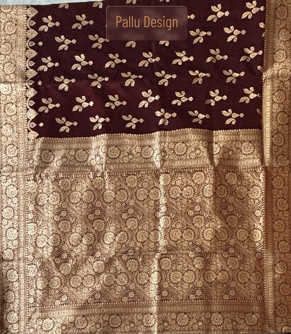 A close-up of a traditional Indian Banarasi Silk saree  with a deep maroon body featuring small gold floral motifs, and a wide ornate gold zari border and pallu section. A semi-transparent label reading 'Pallu Design' is visible near the top.