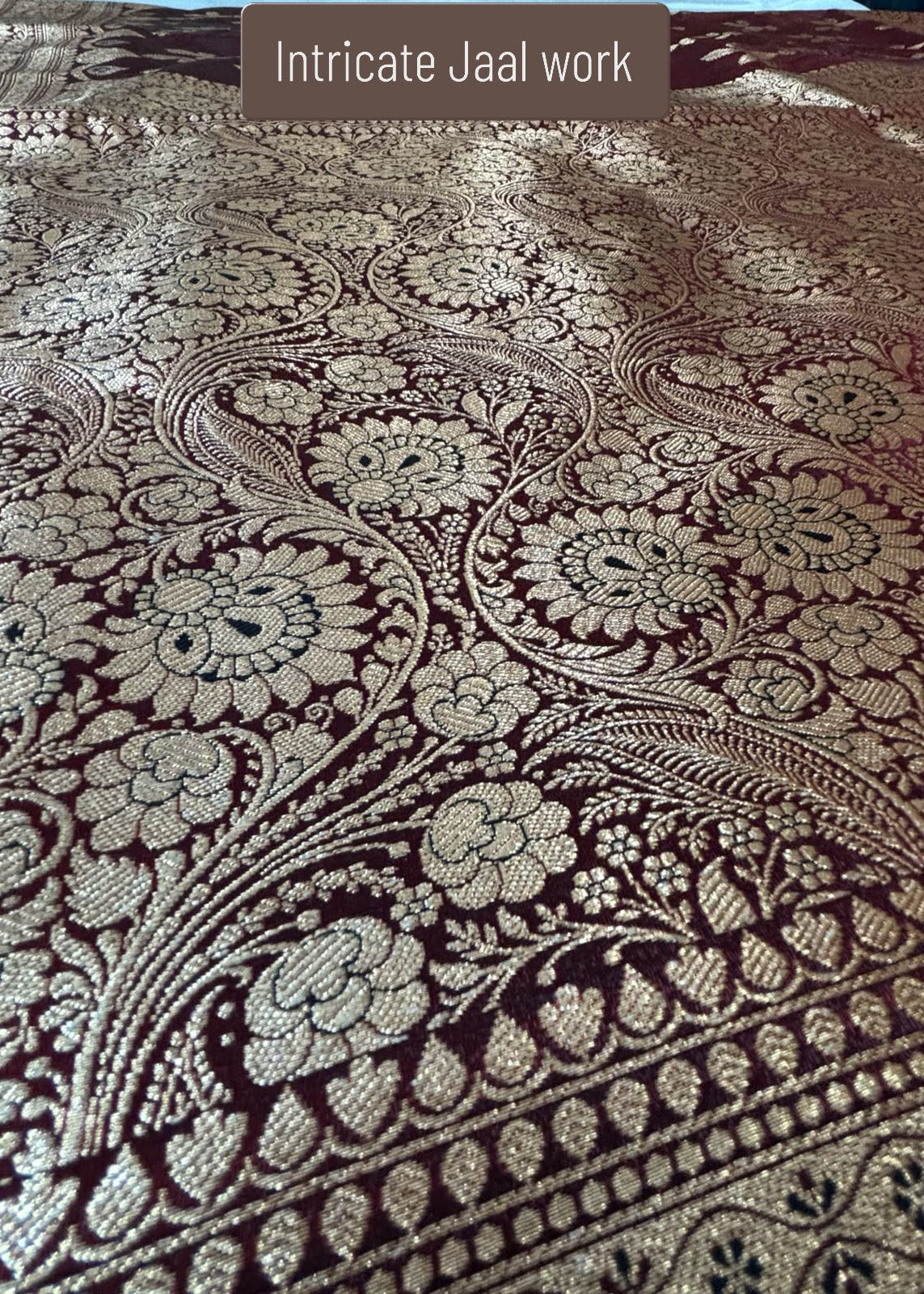 A close-up of Maroon Banarasi Silk Saaree with Golden Zari work featuring intricate Jaal work. A brown label at the top reads 'Intricate Jaal work'.