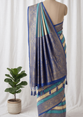 A blue and gold brocade saree with teal stripes and tassel borders, displayed on a mannequin torso against a white curtain backdrop, with a potted green plant on the left.