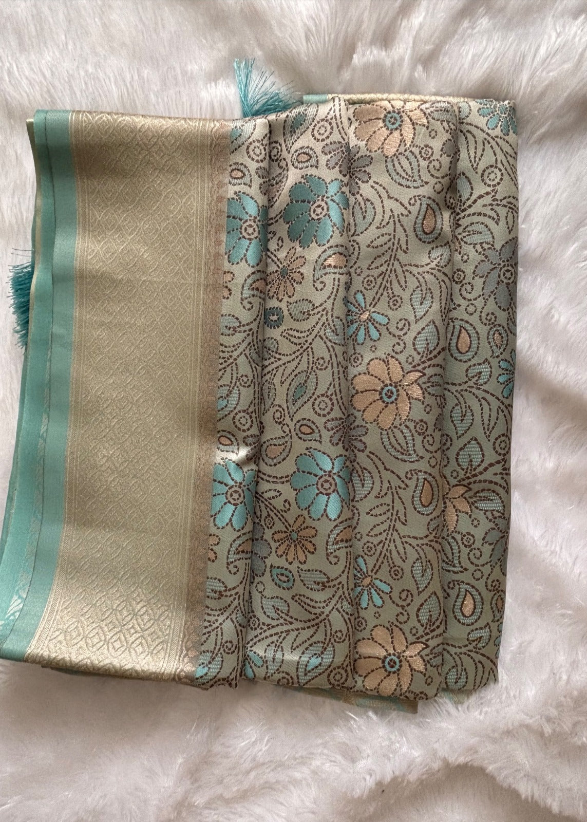 A folded patterned Banarasi Saree, featuring a beige and gold brocade border with teal tassels and a central floral print in teal and peach on a beige background, laid flat on a white faux fur surface.