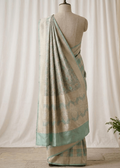 A draped Banarasi saree in beige and mint green with subtle metallic sheen, displayed on a mannequin torso against a cream-colored curtain backdrop with a small potted plant on the side.