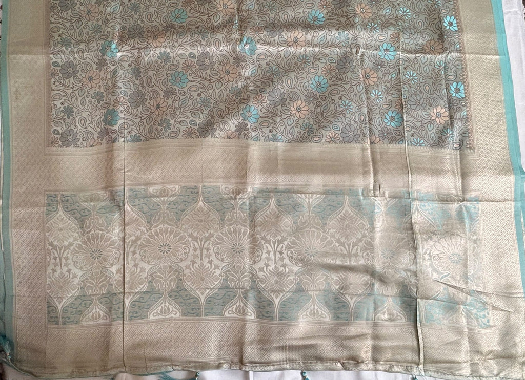 A close-up of a Banarasi Saree, featuring a beige and light blue floral pattern with metallic sheen and decorative borders.