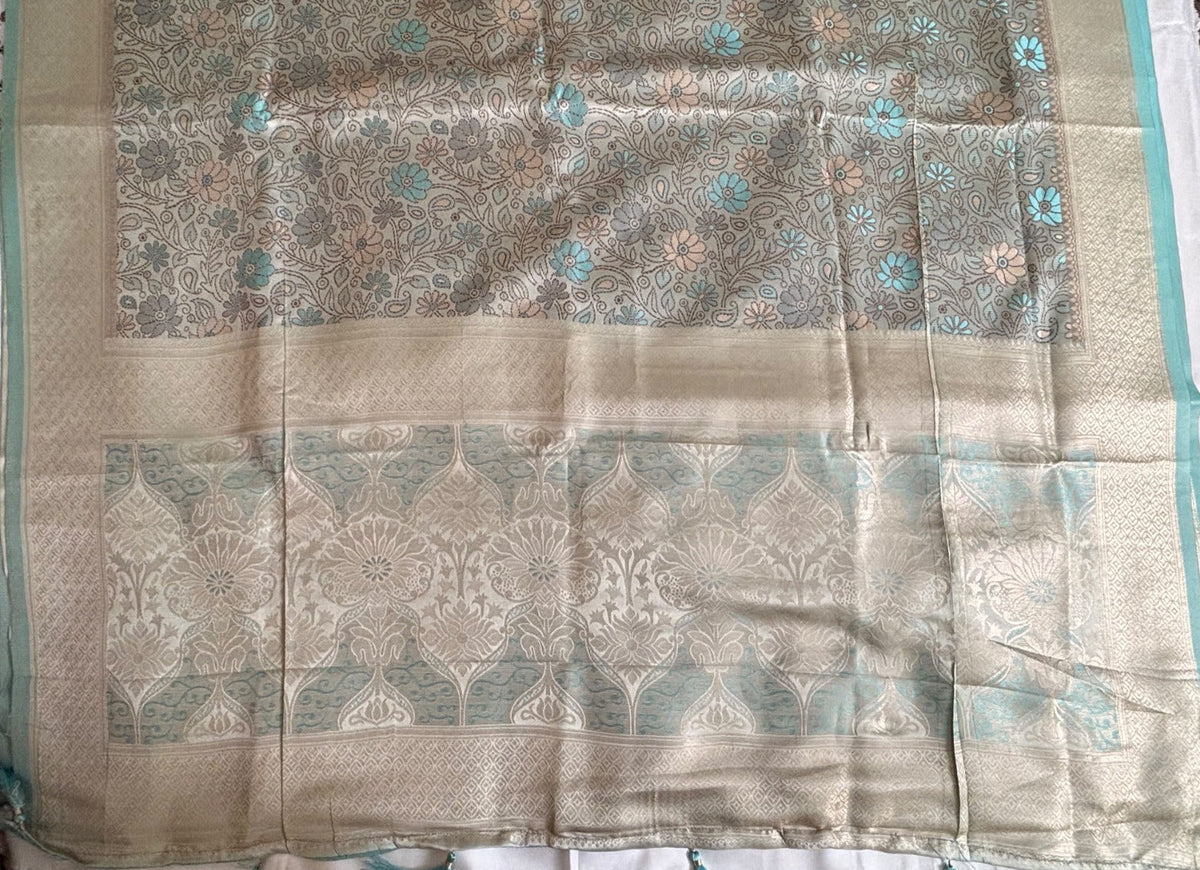 A close-up of a Banarasi Saree, featuring a beige and light blue floral pattern with metallic sheen and decorative borders.