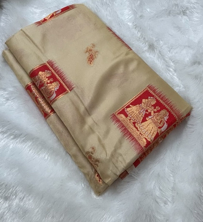 A folded beige silk Baluchari saree with red borders and gold zari motifs, displayed on a white faux fur background.