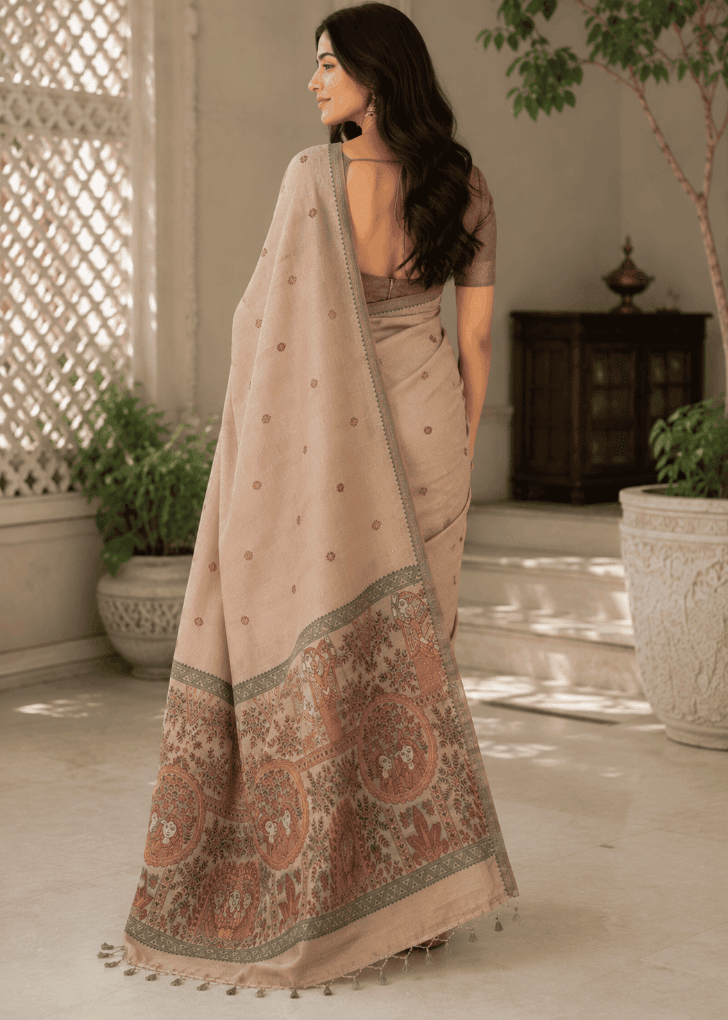 Full-length view of a woman elegantly posing in a lightweight, sandstone-beige Baluchari silk saree. The saree features delicate, small 'buti' motifs on the main body and an intricate, contrasting maroon and beige pallu with traditional mythological figures and narrative scenes. The style is classic and refined, perfect for festive or heritage wear