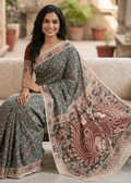 A woman seated on a white sofa wearing a premium teal-green and rust-red cotton Kalamkari saree with a detailed peacock and tree-of-life motif pallu.