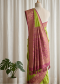 A lime green and dusty pink saree with gold zari embroidery draped on a cream-colored mannequin, set against a soft white curtain backdrop with a potted plant in the corner.