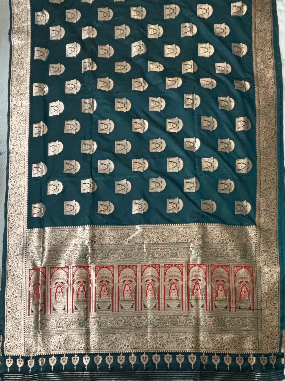 A dark teal green Banarasi silk saree with intricate gold zari motifs, a wide gold border, and red and gold arch motifs along the pallu.