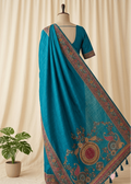 A teal blue saree with intricate multicolor Mandala embroidery and a matching embroidered blouse displayed on a mannequin, set against a cream-colored curtain backdrop with a small potted plant on the left.