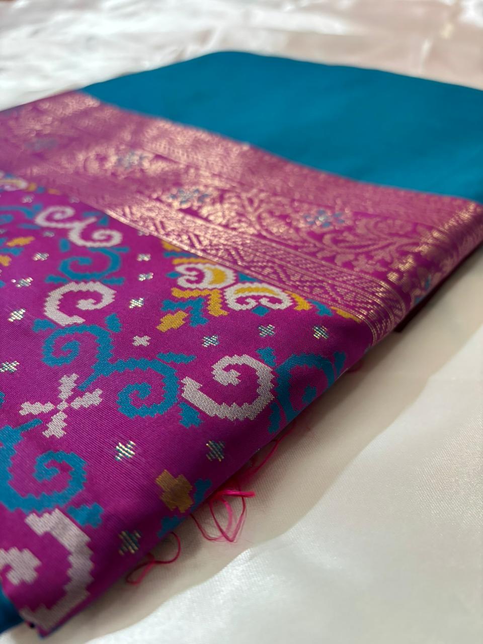 A folded saree of traditional Indian silk fabric, featuring a rich magenta body with intricate gold zari motifs and a contrasting teal blue border.