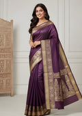 Woman wearing a purple saree with gold border against a white wall.
