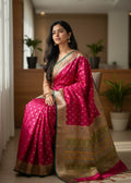 A Woman in a pink Baluchari saree with gold patterns sitting indoors.