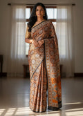 A woman wearing a patterned orange Ajrakh saree is standing in a room with curtains.