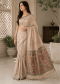 Full-length view of a woman elegantly posing in a lightweight, sandstone-beige Baluchari silk saree. The saree features delicate, small 'buti' motifs on the main body and an intricate, contrasting maroon and beige pallu with traditional mythological figures and narrative scenes. The style is classic and refined, perfect for festive or heritage wear