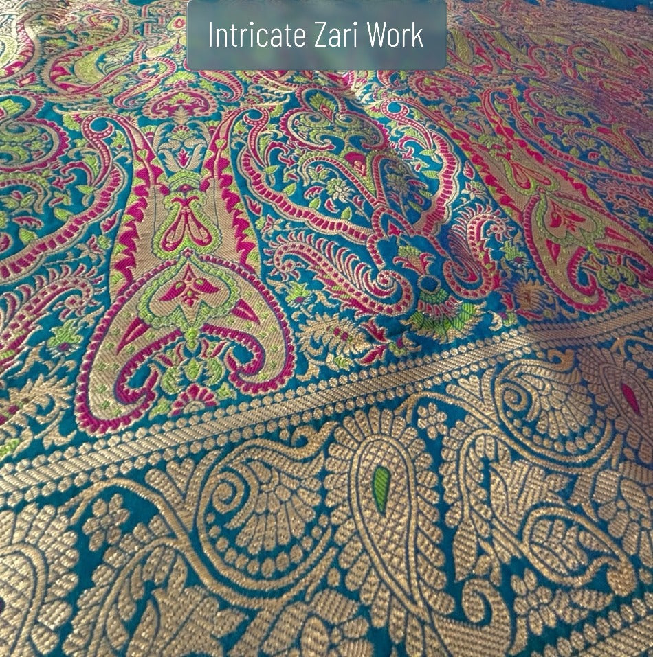 A close-up of a richly patterned zari work fabric with intricate paisley and floral motifs in gold, pink, green, and turquoise blue. A semi-transparent label at the top reads 'Intricate Zari Work'.