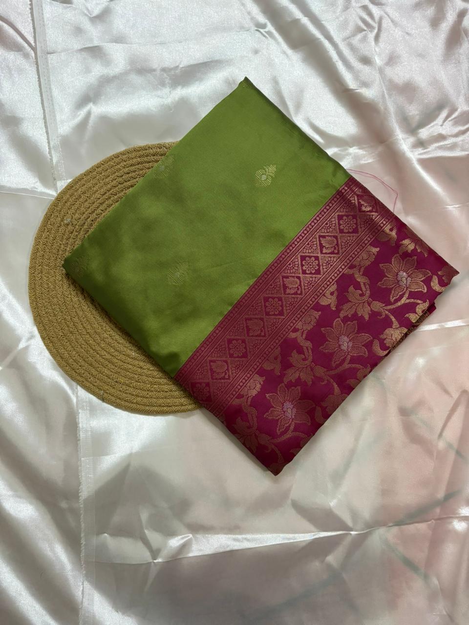 A folded green silk saree with a magenta border featuring gold floral zari work, displayed on a white satin fabric background next to a round woven mat.