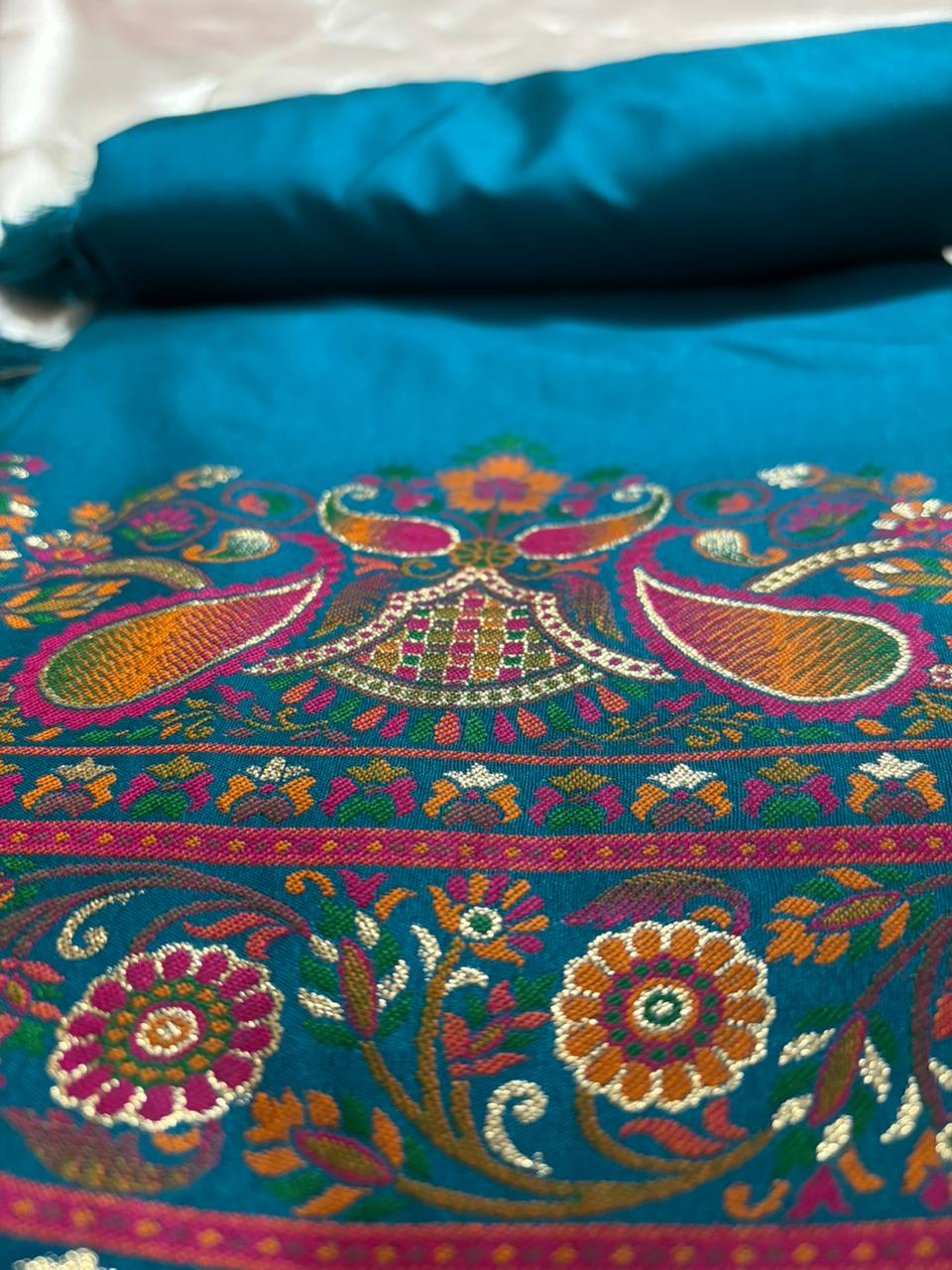 A close-up of a peacock teal-blue saree with intricate multicolor mandala embroidery featuring paisley and floral motifs.