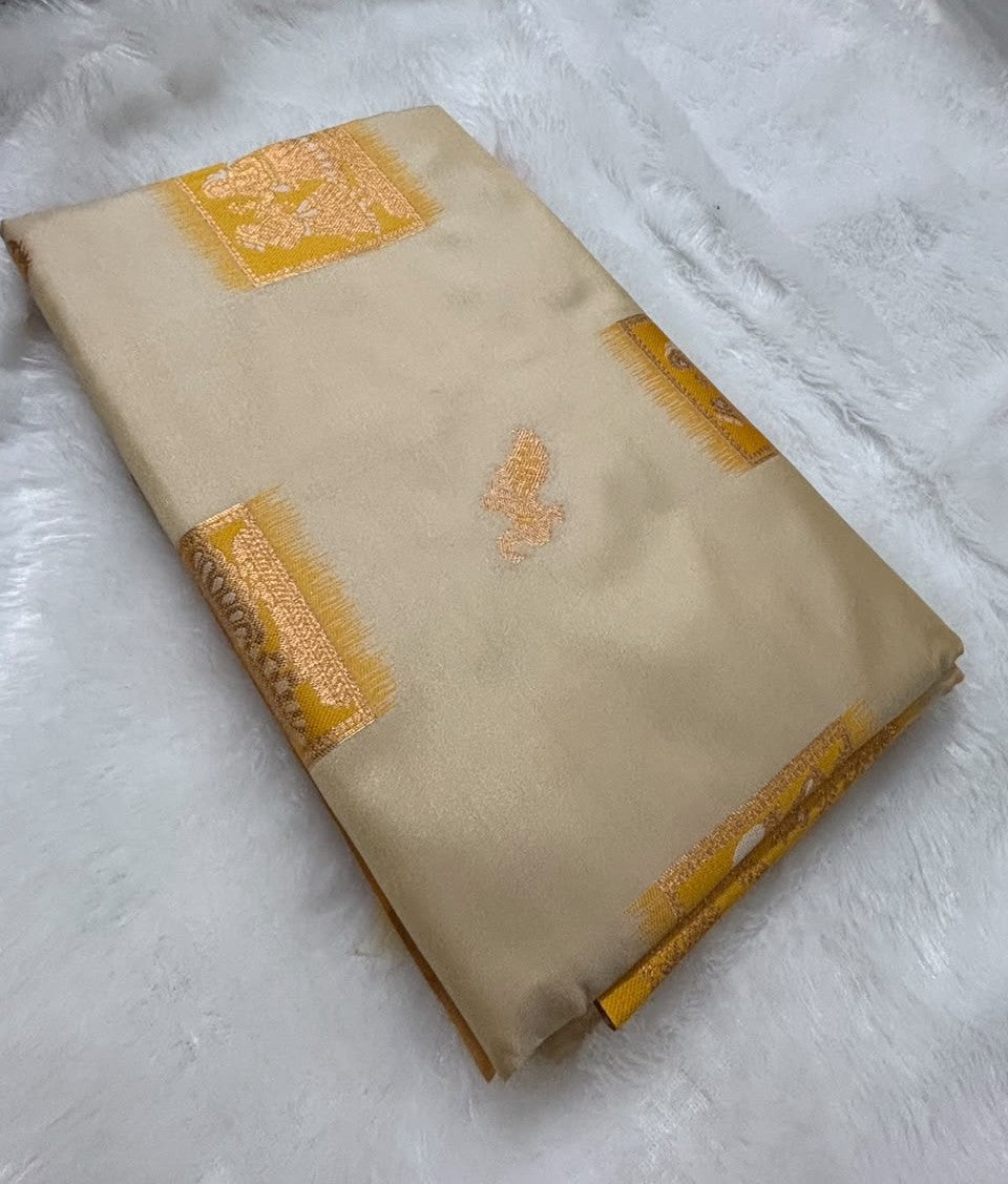 A folded beige Baluchari silk saree with yellow and gold zari woven patterns, displayed on a white faux fur surface.