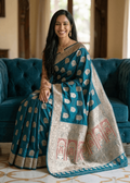 A woman wearing a teal blue and gold Baluchari silk saree sits on a teal velvet sofa in a traditional Indian home. The saree features intricate gold zari motifs and a wide gold border with red and gold patterns.
