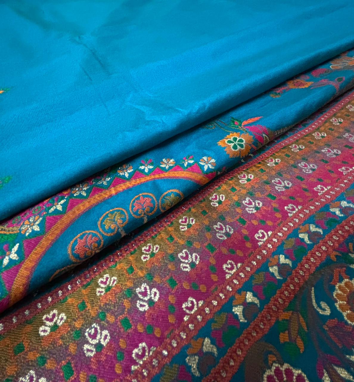 folded view of the peacock teal blue saree  featuring an ornate, multicolored woven border with floral and paisley patterns in pink, orange, yellow, and white.