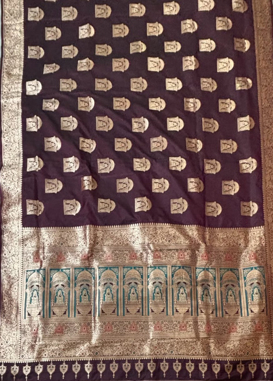 A dark wine-colored Baluchari silk saree with gold zari motifs and a wide gold border, featuring a teal and gold floral patterned pallu.