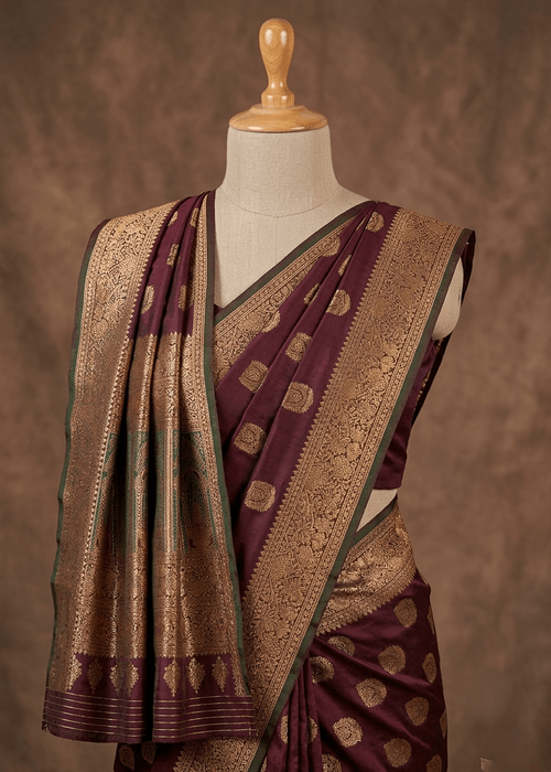 A dark wine colour Baluchari silk saree with intricate zari embroidery, displayed on a mannequin torso against a textured brown backdrop.